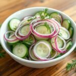 A beautifully arranged cucumber salad in a white bowl with red onions and fresh dill