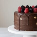 Elegant chocolate drip cake with gold accents and fresh berries on a marble cake stand